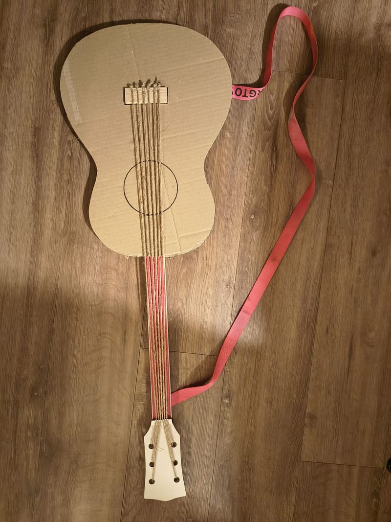 Zelfgemaakte akoestische gitaar van karton met touwtjes als snaren en een roze draagband.