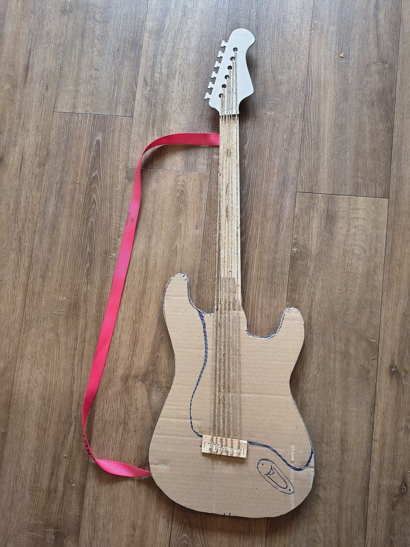 Zelfgemaakte elektrische gitaar van karton met een witte kop en rode draagband.